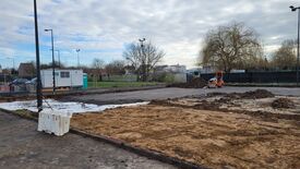 L’image montre un chantier avec une large zone de terre, dont certaines parties semblent avoir été récemment excavées ou nivelées. À l’arrière-plan, on aperçoit un bâtiment modulaire blanc et une toilette mobile verte. Sur la droite, un petit engin de chantier orange, probablement un compacteur, est visible. Le site est entouré de clôtures, et plusieurs arbres ainsi que des bâtiments résidentiels se trouvent en arrière-plan. Le ciel est partiellement couvert de nuages.