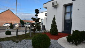 L’image montre un jardin paysager situé devant un bâtiment en briques blanches. Le jardin est composé de différentes zones décoratives : graviers blancs, paillis rouge et pierres grises. On y trouve divers végétaux, dont des buissons taillés et des arbres. Un climatiseur est fixé au mur du bâtiment. À l’arrière-plan, on aperçoit des voitures garées et un autre bâtiment en briques rouges. Le ciel est partiellement nuageux.
