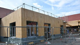 Un bâtiment est en cours de construction ou de rénovation. Sa façade est en bois clair, avec des fenêtres encadrées de métal. Des barrières de sécurité métalliques et une clôture temporaire entourent le bâtiment pour restreindre l’accès. Une échelle métallique est appuyée contre le côté droit du bâtiment, menant au toit. Une clôture de sécurité est également visible sur le toit, probablement pour protéger les ouvriers. Le ciel est partiellement nuageux, laissant apparaître quelques zones bleues.