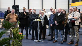 rassemblement. Les participants portent des tenues décontractées ou semi-formelles. Au centre du groupe se trouve une table haute décorée de fleurs. Des plantes en pot sont visibles au premier plan. En arrière-plan, on distingue des éléments industriels comme de grandes portes et un système de sonorisation.