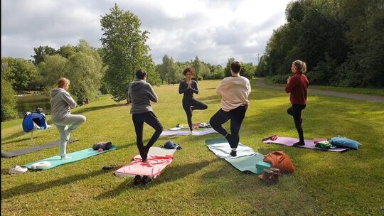L’image montre un groupe de cinq personnes effectuant une séance de yoga en extérieur, sur une pelouse. Chacune est debout sur un tapis de yoga, en posture de l’arbre : une jambe levée et les mains jointes devant la poitrine. L’environnement est verdoyant, avec des arbres et de la végétation en arrière-plan, évoquant un cadre paisible de type parc.