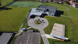 L’image montre une vue aérienne d’un complexe sportif. Deux grands terrains de football sont visibles : l’un en herbe naturelle, l’autre en gazon synthétique. Au centre, un bâtiment est entouré d’une allée circulaire avec des voitures garées. Un parking se trouve à proximité immédiate. À droite du bâtiment, plusieurs surfaces en béton semblent destinées à des activités sportives variées. L’ensemble est entouré de champs verts et de maisons résidentielles en arrière-plan.