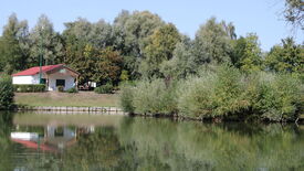 Scène paisible au bord d’un lac avec un petit bâtiment aux murs blancs et au toit rouge, situé en bordure de l’eau. Le bâtiment est entouré de verdure et d’arbres. L’eau calme du lac reflète le bâtiment et la végétation environnante. Le ciel est dégagé, suggérant une météo favorable. Aucune personne ni animal n’est visible.