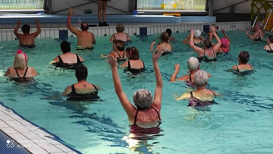 L’image montre un groupe de personnes prenant part à une séance d’aquagym dans une piscine couverte. Les participants sont debout dans l’eau, les bras levés, suivant les mouvements d’un instructeur situé sur le bord du bassin. La profondeur de l’eau est indiquée comme étant de 1,05 mètre. L’environnement est lumineux et adapté à une activité sportive encadrée.