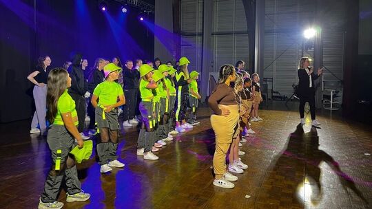 L’image montre un groupe d’enfants et d’adolescents debout en lignes sur une scène, probablement en préparation ou en pleine représentation. Deux groupes distincts sont visibles : l’un porte des hauts verts vifs avec des pantalons noirs et des chapeaux, l’autre des hauts bruns avec des pantalons beiges. Sur le côté droit de la scène, un adulte parle dans un micro, semblant donner des instructions ou présenter le spectacle. L’éclairage de la scène est coloré, contribuant à une ambiance festive.