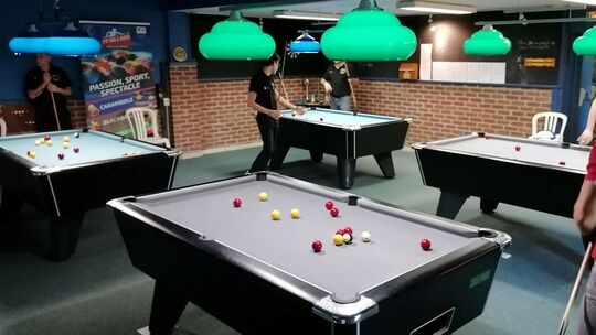 L’image montre une salle de billard intérieure avec quatre tables de jeu. Plusieurs personnes sont en train de jouer ou se préparent à jouer. Chaque table est éclairée par une lampe suspendue de couleur verte. En arrière-plan, un mur en briques est visible, ainsi qu’une affiche portant les mots « PASSION, SPORT, SPECTACLE », « CARAMBOLE », « BLACKBALL » et d’autres textes partiellement masqués.