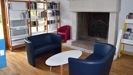 L’image représente un coin lecture chaleureux dans une bibliothèque ou une salle d’étude. Trois fauteuils — deux bleus et un rouge — sont disposés autour d’une petite table basse ovale blanche. En arrière-plan, une grande cheminée avec un couvercle décoratif en métal noir occupe le centre du mur. À gauche de la cheminée, des étagères sont remplies de livres, tandis qu’à droite, un chariot jaune contient d’autres livres et magazines. Le sol est en parquet bois, ce qui renforce l’ambiance accueillante de la pièce.