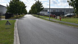 L’image montre une route droite et vide, bordée d’arbres de chaque côté. Sur la droite, un bâtiment porte l’inscription "POLE ARTISANAL BRUAY CO", avec plusieurs voitures stationnées dans un espace clôturé adjacent. Un panneau de signalisation triangulaire est visible sur la bande herbeuse à gauche de la route, et une borne incendie se trouve sur le trottoir à droite. Le ciel est partiellement nuageux. Aucun piéton ni véhicule en mouvement n’est visible.