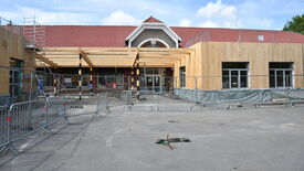 Un bâtiment est en cours de construction ou de rénovation. Il possède un toit en tuiles rouges et une ossature en bois est en cours d’installation sur la façade avant. Des barrières métalliques entourent la zone de travaux. Une échelle est visible sur le côté gauche de l’image. Le sol devant le bâtiment est pavé, et une trottinette électrique est couchée au sol au premier plan.