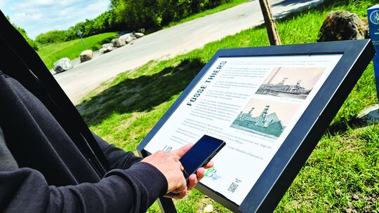 L’image montre une personne tenant un smartphone, en train de lire un panneau d’information situé en extérieur. Le panneau est intitulé "FOSSE THIERS" et contient du texte explicatif ainsi que des images ou illustrations liées au sujet.

L’environnement autour du panneau est naturel : on y voit de la végétation, un chemin, des rochers et des arbres, suggérant un site de promenade ou un lieu patrimonial.