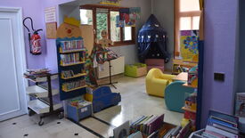 Médiathèque photo 08 Espace de lecture pour enfants dans une bibliothèque ou un lieu éducatif. Des étagères colorées, certaines en forme de personnages ou objets ludiques (comme une voiture ou un clown), sont remplies de livres pour enfants. À gauche, un extincteur est fixé au mur, à côté d’un petit chariot contenant d’autres livres. En arrière-plan, un coin lecture cosy est décoré de rideaux bleu foncé ornés d’étoiles et de lunes, avec des assises moelleuses aux couleurs vives (jaune, bleu). De grandes fenêtres laissent entrer la lumière naturelle. Des affiches murales promeuvent la lecture pour les enfants.