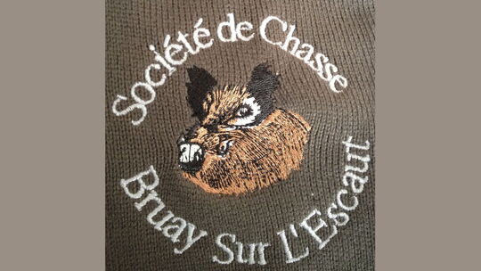 L’image montre une broderie sur tissu représentant la tête d’un sanglier au centre. Autour de cette tête, un texte en arc de cercle est inscrit : « Société de Chasse » en haut et « Bruay Sur L’Escaut » en bas. Le style et la disposition suggèrent un écusson ou un emblème officiel, probablement utilisé par une association de chasse locale.