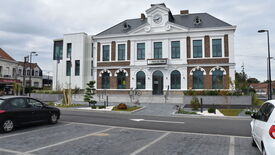 L’image montre un bâtiment de style classique portant l’inscription « Hôtel de Ville », indiquant qu’il s’agit d’une mairie. La façade est en briques avec des éléments blancs, des fenêtres en arc au rez-de-chaussée et rectangulaires à l’étage. Une horloge est centrée au sommet du bâtiment. Des escaliers mènent à l’entrée principale, entourée de petits arbres et plantes décoratives. Deux vélos sont stationnés près de l’entrée, et plusieurs voitures sont garées sur la rue pavée devant le bâtiment.