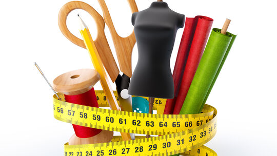 L’image présente un ensemble d’outils de couture et de tailleur. Au centre, un mannequin noir de couture est entouré de grands ciseaux en bois, d’un crayon jaune avec gomme, d’une bobine de fil rouge, d’une aiguille enfilée, de deux rouleaux de tissu (rouge et vert), ainsi que d’un mètre ruban jaune enroulé autour des objets. L’ensemble évoque un espace de travail dédié à la confection textile.