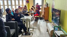 Un groupe de personnes se trouve dans une pièce où se déroulent diverses activités. À gauche, deux personnes sont assises : l’une tient une manette de jeu, l’autre utilise un volant connecté à une console. À l’arrière-plan, plusieurs personnes sont rassemblées autour d’une table avec des ordinateurs portables et d’autres appareils. Sur la droite, un grand écran de télévision affiche ce qui semble être un jeu de course. La pièce est éclairée par la lumière naturelle provenant de grandes fenêtres. Une décoration murale représente Spider-Man suspendu au plafond.