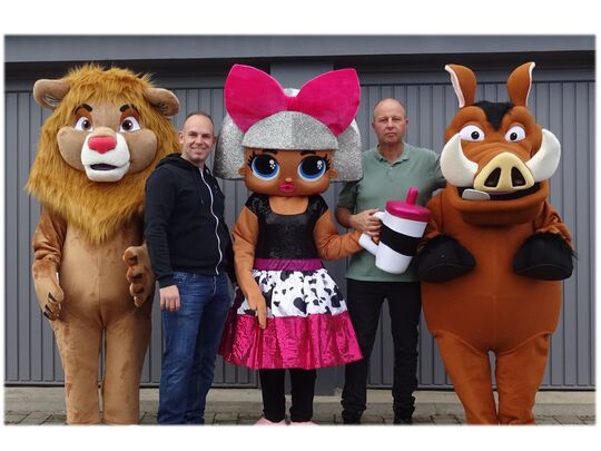 L’image montre cinq personnes debout côte à côte. Deux d’entre elles portent des vêtements décontractés, tandis que les trois autres sont déguisées dans des costumes volumineux et colorés. À gauche, un costume de lion ; au centre, un personnage avec de longs cheveux argentés, un grand nœud rose et tenant une tasse ; à droite, un costume de phacochère. Les visages des deux personnes en tenue décontractée sont floutés pour préserver leur anonymat. L’arrière-plan est constitué d’une grande porte grise, semblable à une porte de garage.