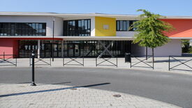 Bâtiment moderne avec une façade colorée composée de sections rouges, jaunes et orange. Il possède de grandes fenêtres et un toit plat. À droite, un arbre est visible. Au premier plan, un rond-point avec des barrières métalliques et des lampadaires se trouve devant le bâtiment.