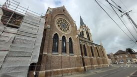 L’image montre une grande église en briques de style gothique, avec des arcs en ogive et une haute flèche. La façade est ornée d’une grande rosace en vitrail, située au-dessus de trois fenêtres étroites et élancées. Un échafaudage est visible sur le côté gauche du bâtiment, indiquant des travaux en cours. Des bâtiments résidentiels et des lignes électriques entourent l’église.