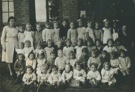 Photographie de classe montrant un groupe d’enfants accompagnés de deux adultes, posant devant un bâtiment en briques. Les enfants sont disposés en plusieurs rangées : certains sont assis au sol, d’autres debout derrière.
