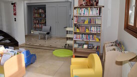 L’image montre un petit espace de lecture, probablement destiné aux enfants. Plusieurs étagères sont remplies de livres. Sur la gauche, une étagère en forme de voiture bleue attire l’attention. Au premier plan, un fauteuil jaune est placé près d’un tapis rond vert. À l’arrière-plan, une autre étagère contient davantage de livres, accompagnée d’un fauteuil gris. Le sol est carrelé, les murs sont blancs, et une fenêtre sur la droite laisse entrer la lumière naturelle, rendant l’espace lumineux et accueillant.