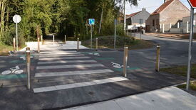 Zone de traversée pour piétons et cyclistes avec un passage piéton zébré et des voies cyclables de chaque côté, marquées par des symboles de vélo au sol. Le chemin mène vers une zone boisée, tandis que l’autre extrémité rejoint une route bordée de maisons. Plusieurs bornes en bois empêchent l’accès des véhicules. Des panneaux de signalisation indiquent les chemins pour piétons et cyclistes.