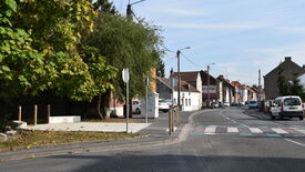 Rue calme dans un quartier résidentiel, bordée de maisons de chaque côté. Plusieurs voitures sont stationnées le long de la route, et d’autres circulent. À gauche, un grand arbre feuillu se dresse près d’une petite zone pavée avec des bornes. La route, marquée d’un passage piéton, tourne vers la droite. Des poteaux électriques avec des câbles longent la rue.