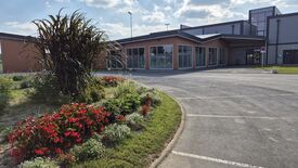 Un bâtiment moderne avec de grandes fenêtres vitrées est visible, avec un parking situé à l’avant. Sur la gauche de l’image, une zone paysagée est aménagée avec diverses plantes et fleurs, dont des fleurs rouges et des herbes hautes. Le ciel est partiellement nuageux.