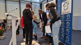 Un groupe composé d’enfants et d’adultes (dont Madame La Maire) est rassemblé sur une scène ou une estrade. Les enfants portent des tenues de sport, avec le numéro 4 visible sur l’un d’eux. Les adultes semblent remettre des objets ou des récompenses aux enfants. À gauche, un pupitre avec un micro est visible, et à droite, une bannière affiche le logo et le nom « Région Hauts-de-France » à plusieurs reprises. L’événement se déroule en intérieur, dans un espace lumineux avec de grandes fenêtres à l’arrière-plan.