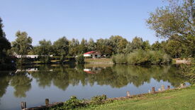 Paysage paisible avec un lac calme reflétant les arbres environnants et une petite maison au toit rouge. La maison est située de l’autre côté du lac, entourée de verdure et d’arbres. Au premier plan, de l’herbe et des poteaux en bois bordent la rive. Le ciel est bleu et dégagé, suggérant une météo clémente.