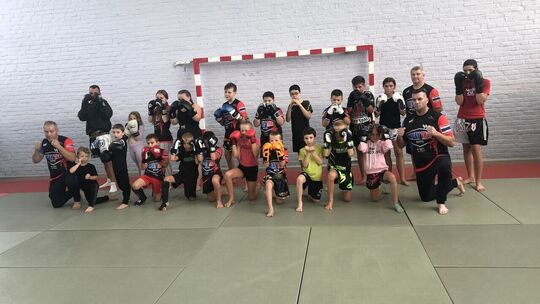 L’image montre un groupe composé d’enfants et d’adultes posant dans une salle de sport dédiée aux arts martiaux. Tous portent des gants de boxe et des tenues de sport de combat. Certains sont debout, d’autres agenouillés sur des tapis. En arrière-plan, on distingue un mur en briques blanches et un but de handball ou de futsal.
