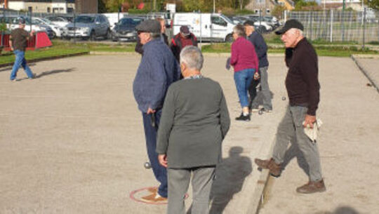 L’image montre un groupe de personnes debout ou marchant sur une surface plane et sablonneuse, probablement un terrain de pétanque. Plusieurs individus tiennent des boules dans les mains, suggérant qu’ils participent à une partie. À l’arrière-plan, on aperçoit des voitures garées et des bâtiments, indiquant un environnement urbain ou périurbain.