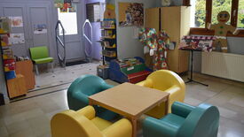Médiathèque photo 09 Espace de lecture pour enfants dans une bibliothèque. Au centre, des chaises colorées entourent une petite table. Des étagères remplies de livres bordent la pièce. La décoration comprend un grand personnage en pain d’épices, un arbre fait de cercles en papier, ainsi que d’autres œuvres artistiques accrochées aux murs. De grandes fenêtres laissent entrer la lumière naturelle.