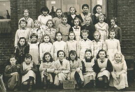Photographie de groupe représentant des enfants, probablement une photo de classe, posant devant un bâtiment en briques. Les enfants sont disposés en plusieurs rangées, certains assis, d’autres debout. Les visages ont été floutés pour préserver leur anonymat. Un panneau placé au centre en bas de l’image porte l’inscription : « 4E 1952–1953 »
