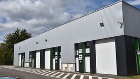 L’image montre un bâtiment industriel moderne avec un revêtement en tôle ondulée, peint en gris, noir et vert. Trois portes de garage sont visibles, dont deux sont étiquetées "BRUAY CO 13" et "BRUAY CO 14". Devant le bâtiment, on distingue des places de stationnement marquées au sol ainsi qu’un passage piéton rayé. À l’arrière-plan, des arbres indiquent la présence de verdure autour du site.