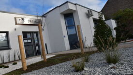 L’image montre l’extérieur d’un bâtiment en briques blanches avec de grandes fenêtres. Une enseigne au-dessus de l’entrée indique "Pôle Social". Un climatiseur est fixé sur le mur. Devant le bâtiment, un espace paysager est aménagé avec du gravier, de petites plantes et des poteaux en bois.