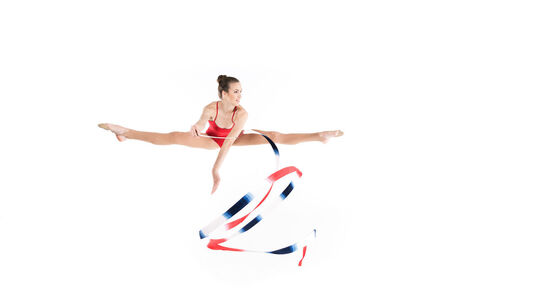 L’image montre une gymnaste en plein saut, réalisant un grand écart en l’air tout en tenant un ruban de gymnastique rythmique. Elle porte un justaucorps rouge, et le ruban qu’elle manie est aux couleurs rouge, blanc et bleu. Le fond est entièrement blanc, mettant en valeur la posture dynamique et la fluidité du mouvement.