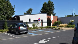 L’image montre un petit parking situé devant un bâtiment moderne aux murs en briques blanches et rouges. Deux places de stationnement réservées aux personnes handicapées sont clairement marquées au sol avec de la peinture bleue et des symboles. Une voiture noire est garée sur une place standard. Un panneau "Stationnement interdit" est visible près de l’entrée du bâtiment, qui est entourée de verdure et de fleurs. Le ciel est bleu et dégagé, suggérant une journée ensoleillée.