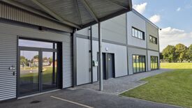 L’image montre un bâtiment contemporain à deux niveaux, habillé d’un revêtement en métal ondulé gris. Sur la gauche, l’entrée principale est constituée de grandes portes vitrées, accompagnées d’un interphone et protégées par un auvent métallique. Au centre de la façade, une porte grise est surmontée d’un panneau jaune, avec un défibrillateur mural vert installé à proximité. Sur la droite, plusieurs fenêtres rectangulaires sont alignées sur les deux étages. Le bâtiment est entouré d’une pelouse verte bien entretenue, sous un ciel bleu parsemé de quelques nuages blancs.