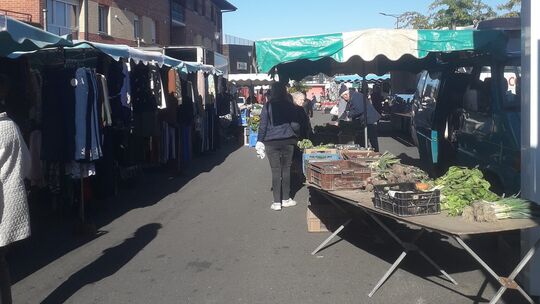 L’image représente un marché en plein air animé. Des stands sont installés sous des auvents verts et blancs. À gauche, des vêtements sont exposés, tandis qu’à droite, une table présente des légumes frais, notamment des légumes-feuilles et des racines. Plusieurs personnes déambulent entre les étals, observant les produits proposés. En arrière-plan, on distingue d’autres stands ainsi qu’un bâtiment en briques.