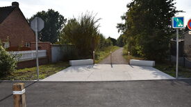 Entrée d’un chemin avec des barrières en béton et des poteaux en bois. Deux panneaux de signalisation sont visibles à droite : l’un indique un chemin pour piétons et cyclistes, l’autre interdit l’accès aux véhicules. À gauche, une maison avec une clôture blanche borde le chemin. Le sentier se prolonge vers une zone boisée à l’arrière-plan.