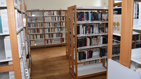 L’image montre l’intérieur d’une bibliothèque avec plusieurs rayonnages en bois bien remplis de livres. Les étagères sont organisées et étiquetées, notamment une section intitulée "POLICIERS" dédiée aux romans policiers, et une autre indiquant les lettres "C" et "D à G" pour le classement alphabétique. Le sol est en parquet bois, et l’espace est bien éclairé, offrant un environnement ordonné et propice à la lecture.