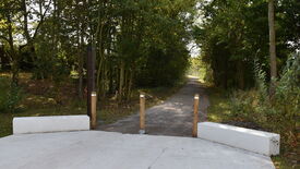 Chemin non goudronné menant dans une zone boisée, encadré à l’entrée par deux blocs de béton blancs et trois poteaux en bois. Un panneau vertical en bois est visible à gauche du chemin, mais son texte est partiellement illisible. La végétation dense forme un effet de tunnel naturel autour du sentier.