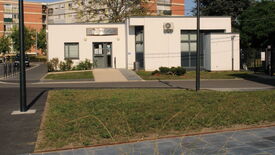 L’image montre un petit bâtiment moderne à façade blanche, doté de grandes fenêtres. Une enseigne au-dessus de l’entrée indique "Pole Social". Un chemin pavé mène à l’entrée, bordé de pelouse et de quelques arbustes. À l’arrière-plan, on aperçoit des immeubles d’habitation plus grands et des arbres. L’environnement semble urbain, avec des trottoirs aménagés et des lampadaires visibles au premier plan.