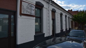 Façade extérieure d’un bâtiment portant l’enseigne « Espace Musical ». Les murs sont en briques blanches avec des encadrements noirs autour des fenêtres et des portes. Plusieurs voitures sont garées devant le bâtiment, et des arbres bordent la rue. Le ciel est partiellement nuageux.
