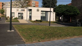 L’image montre un petit bâtiment moderne à façade blanche, doté de grandes fenêtres. Une enseigne au-dessus de l’entrée indique "Pole Social". Un chemin pavé mène à l’entrée, bordé de pelouse et de quelques arbustes. À l’arrière-plan, on aperçoit des immeubles d’habitation plus grands et des arbres. L’environnement semble urbain, avec des trottoirs aménagés et des lampadaires visibles au premier plan.