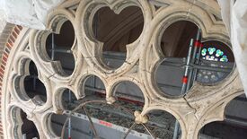 L’image montre un gros plan sur un encadrement de fenêtre en pierre de style gothique, orné de motifs circulaires et d’arcs en ogive. La fenêtre semble en cours de restauration ou de construction, comme en témoignent les échafaudages visibles à l’arrière et une protection partielle dans le coin supérieur gauche. En arrière-plan, un vitrail coloré aux motifs floraux est partiellement visible.