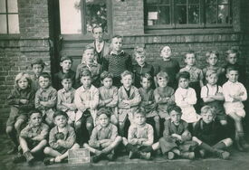 Photographie de groupe représentant des enfants et des adultes posant devant un bâtiment en briques. Le groupe est organisé en trois rangées : la première assise au sol, la deuxième sur des bancs ou des chaises, et la troisième debout. Un petit tableau noir placé devant le groupe porte l’inscription : « Bruay Thiers »