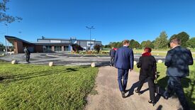Un groupe de personnes (dont Madame La Maire) marche en direction d’un bâtiment moderne, situé derrière un parking. Le bâtiment présente de grandes fenêtres et un aménagement paysager avec des plantes et des fleurs à l’avant. Le ciel est dégagé et bleu, ce qui indique une météo favorable.