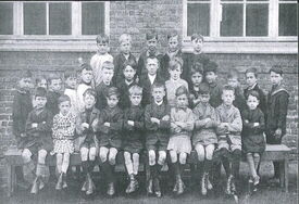 Photographie en noir et blanc représentant un groupe d’enfants posant pour une photo de classe devant un bâtiment en briques avec des fenêtres. Les enfants sont disposés en deux rangées : la première assise sur un banc, la seconde debout derrière. Les vêtements portés évoquent une époque ancienne, probablement du début du XXe siècle.