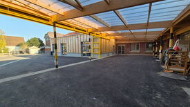 L’image montre un espace extérieur partiellement couvert par une toiture transparente, soutenue par des poutres en bois et des colonnes métalliques jaunes. Sur la droite, on aperçoit des matériaux et équipements de chantier, suggérant un projet de construction ou de rénovation en cours. À l’arrière-plan, des bâtiments aux façades en bois et en briques comportent de grandes fenêtres et des portes. Le sol est asphalté, et l’espace s’ouvre vers une zone non couverte.